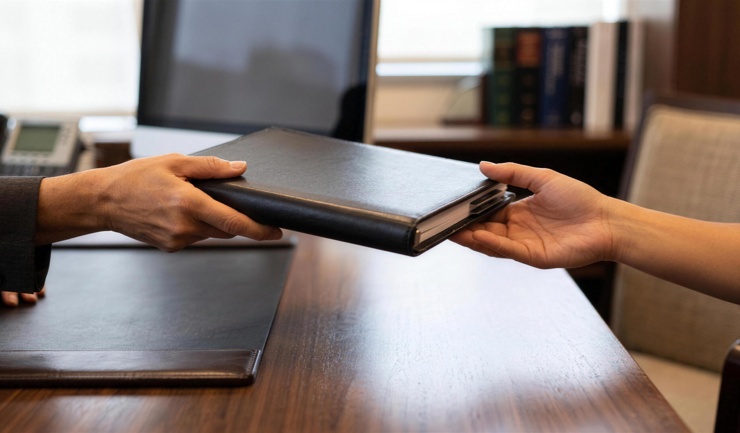 Two hands exchange a leather project binder across an executive office desk.
